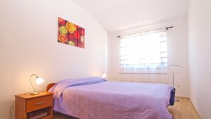 A bedroom with a bed, a nightstand, a lamp, a window with curtains, and a floral painting on the wall. - Apartment 1639 (Pula)