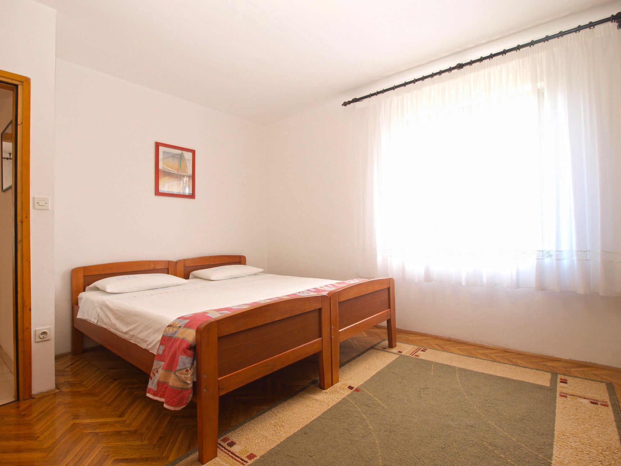 Un dormitorio con una cama de madera, una mesita de noche pequeña, un corredor de cama sencillo y una ventana con cortinas traslúcidas.