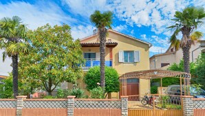 Une maison à deux étages avec un balcon, entourée de palmiers et d’un jardin.