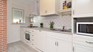 Une cuisine avec des meubles blancs, une cuisinière à gaz, un four à micro-ondes et une fenêtre avec des stores.