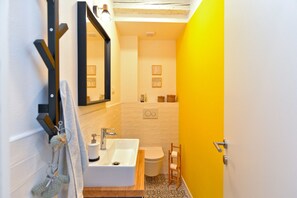Une salle de bain avec un lavabo blanc, un miroir et des toilettes. On y trouve une étagère en bois ornée d’objets décoratifs et un porte-serviettes avec des serviettes.