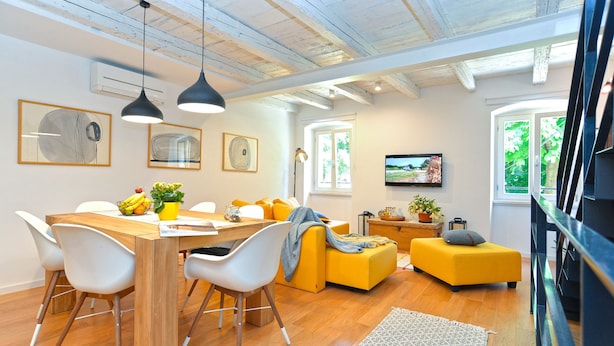 A modern living space with a wooden dining table, white chairs, a yellow sofa, and a TV mounted on the wall. - House 1650 (Vodnjan)