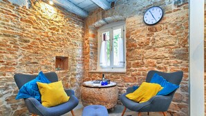 A cozy room with stone walls, two armchairs with cushions, a small round table with a book and a bottle, and a clock on the wall. - House 1650 (Vodnjan)
