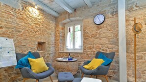 A cozy room with stone walls, two grey armchairs with yellow and blue cushions, a small round table with a bottle and glasses, a wooden floor lamp, and a wall clock. - House 1650 (Vodnjan)