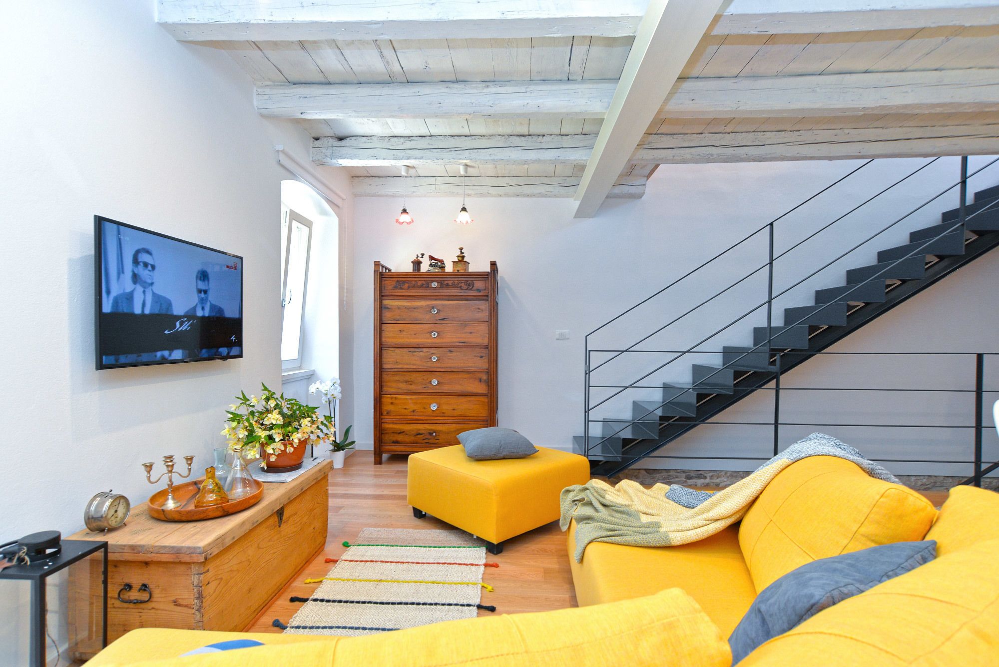 Uma sala de estar moderna com um aparador de madeira, uma TV fixada na parede, uma escada e um sofá amarelo.