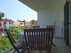 A balcony with a table and two wooden chairs, overlooking a garden and neighboring houses.