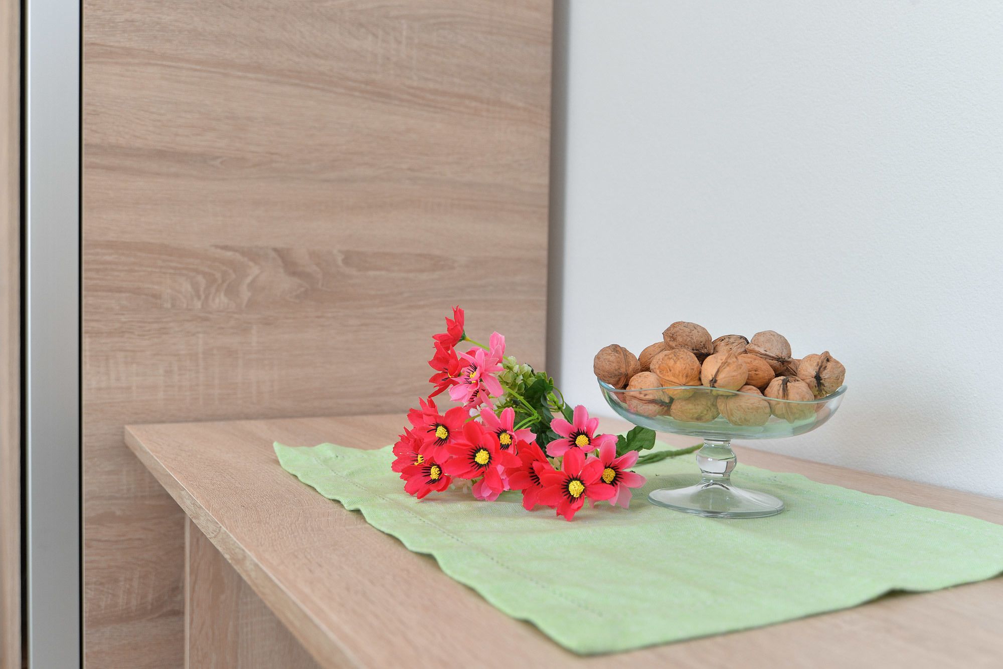 Une table en bois sur laquelle se trouve un saladier en verre rempli de noix et un bouquet de fleurs rouges, le tout posé sur un tissu vert.