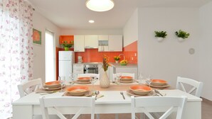 Een eetruimte met een witte tafel voor zes personen, omringd door witte stoelen. Achtergrond is een keuken met oranje en witte kasten, en een koelkast.