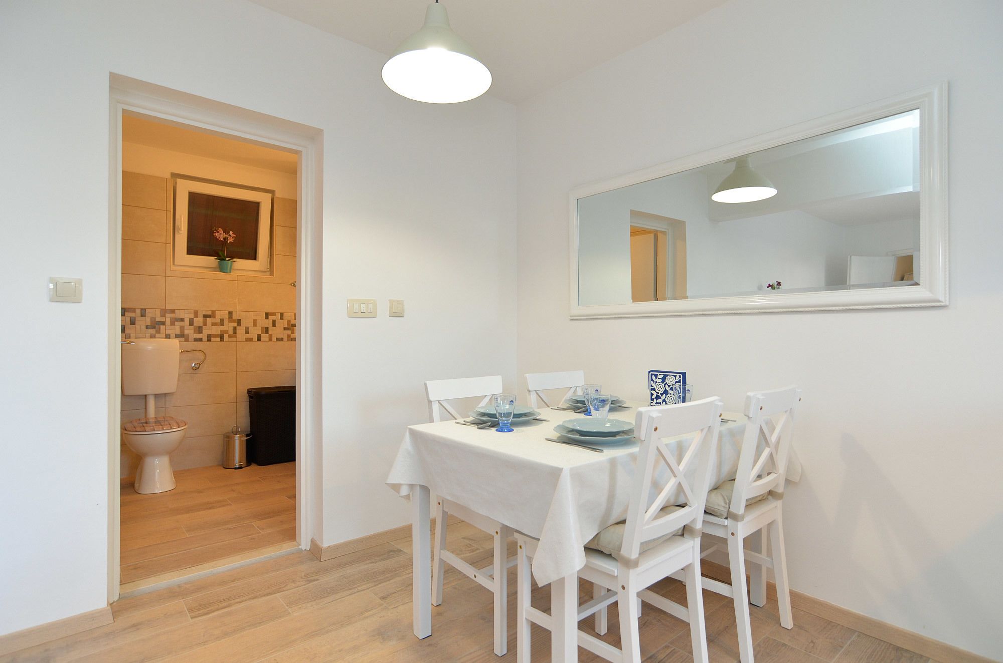 A dining area with a white table set for four, a mirror above the table, and a view into the adjacent bathroom.