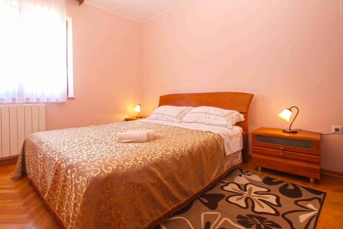 A bedroom with a bed, bedside lamps, a nightstand, a radiator, and a window with curtains.