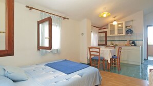 A bedroom with a bed, a window with curtains, a dining area with a table and chairs, and a kitchen with cabinets and a clock. - Apartment 1665 (Vodnjan)