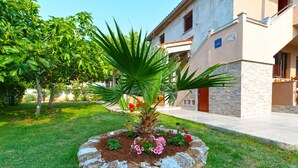 A garden with a palm tree and flowers in front of a building with stone walls and a tiled roof. - Apartment 1665 (Vodnjan)