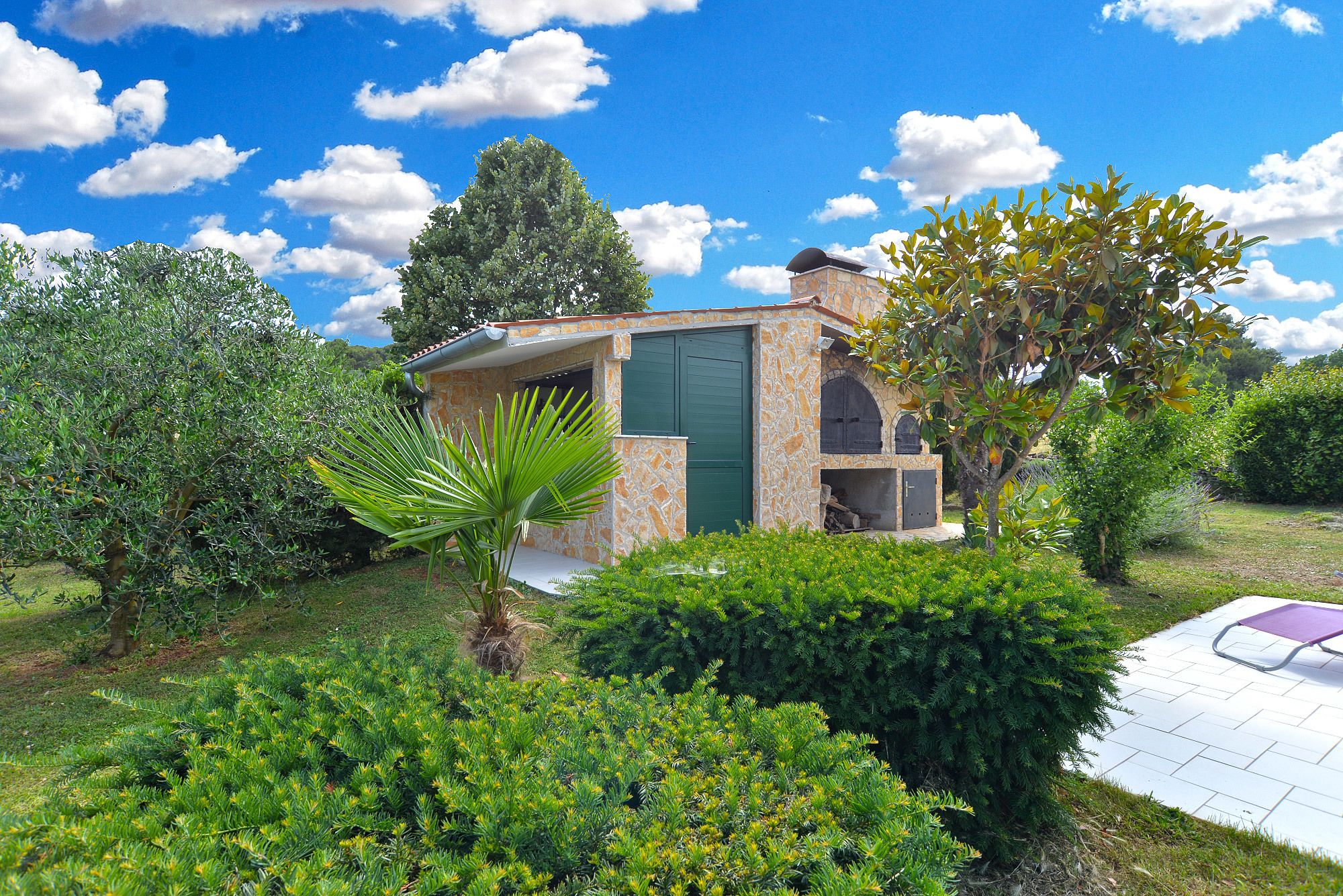 A stone building with a green door, surrounded by lush greenery and a clear blue sky.