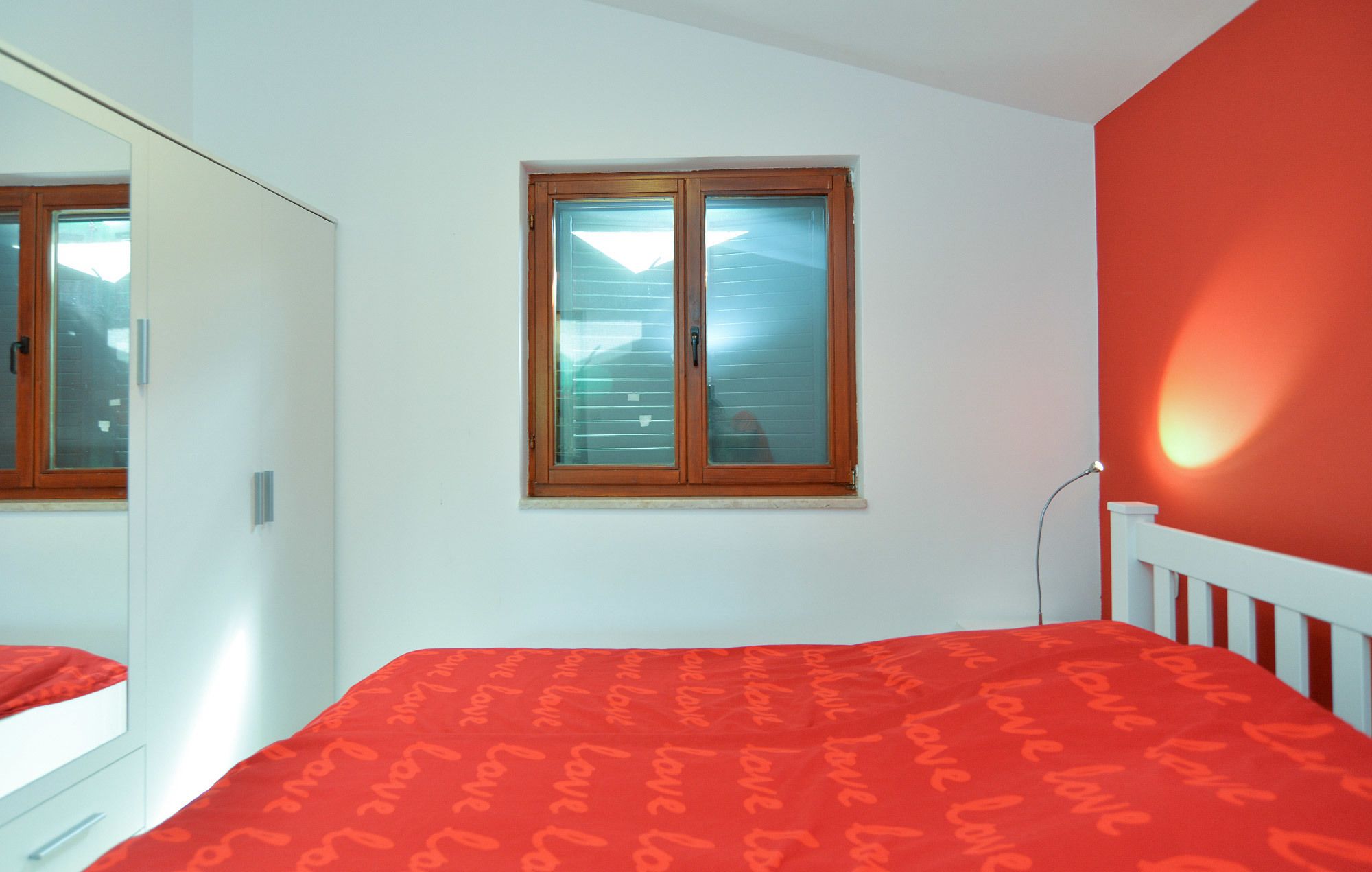 A bedroom with a bed, a wardrobe, a window with blinds, and a red wall.