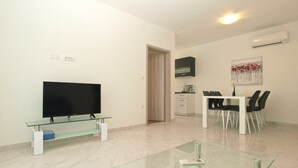 A modern living room with a flat-screen TV on a glass table, a dining area with a white table and black chairs, and a kitchen area with a black cabinet and white appliances. - Apartment 1670 (Fazana)