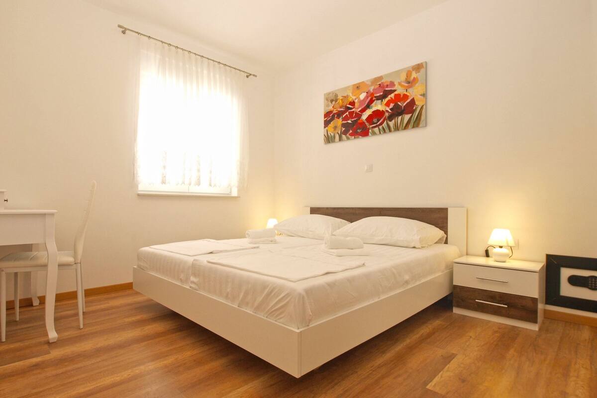Quarto com cama, mesas de cabeceira, uma cadeira, janela com cortinas e um quadro na parede.