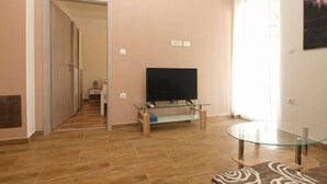 A living room with a flat-screen TV on a glass table, wooden flooring, and a patterned sofa. - Apartment 1670 (Fazana)