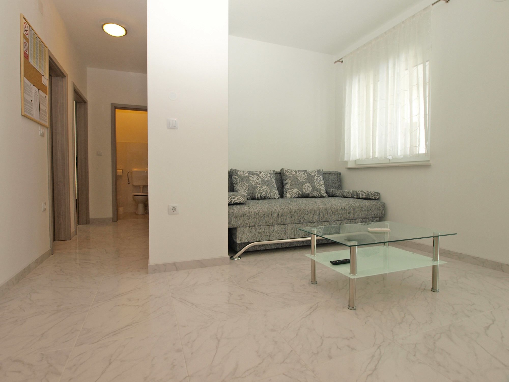 A modern living room with a glass coffee table, grey sofa with patterned cushions, and a window with sheer curtains.