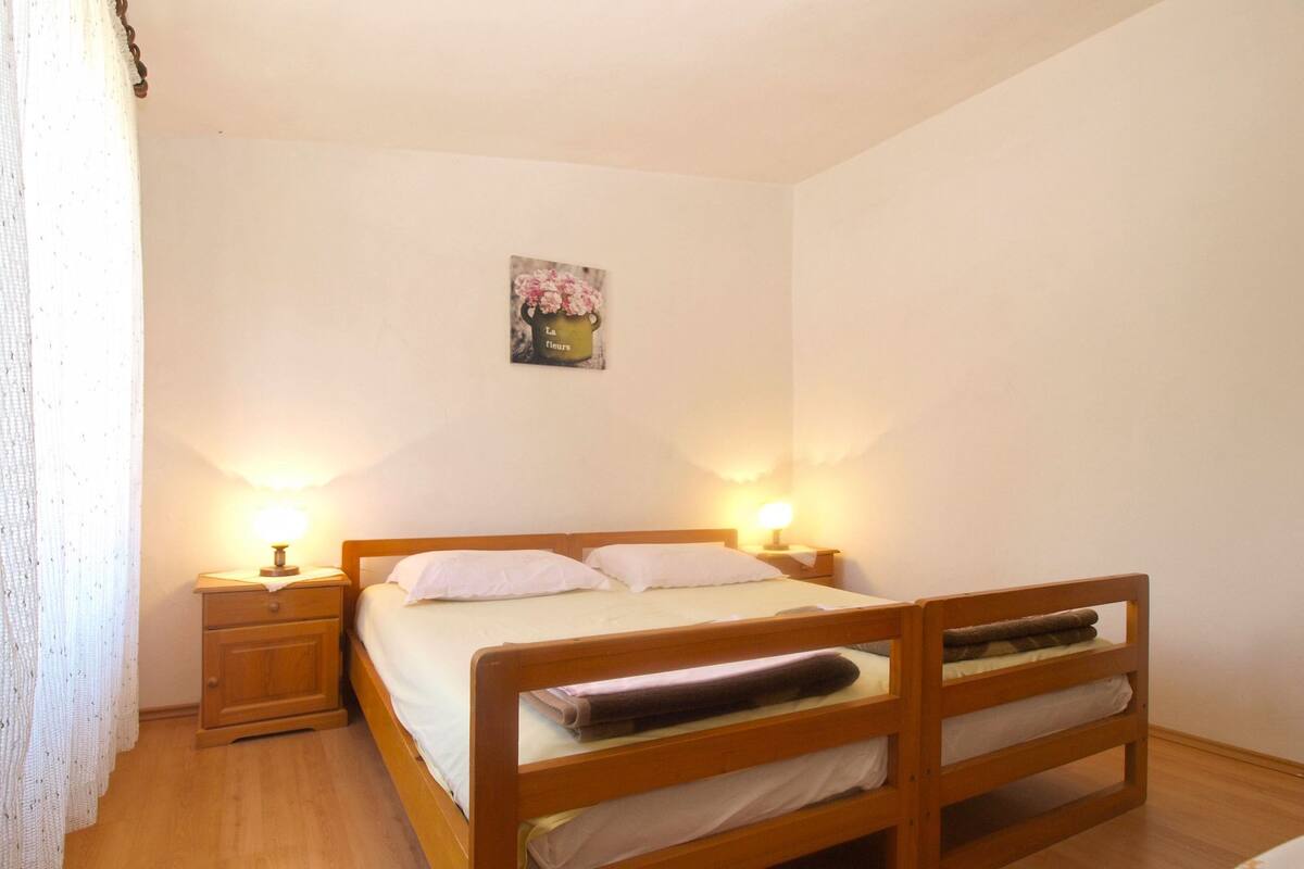 A single bed with a wooden frame, two bedside tables with lamps, a small painting on the wall, and a window with curtains.