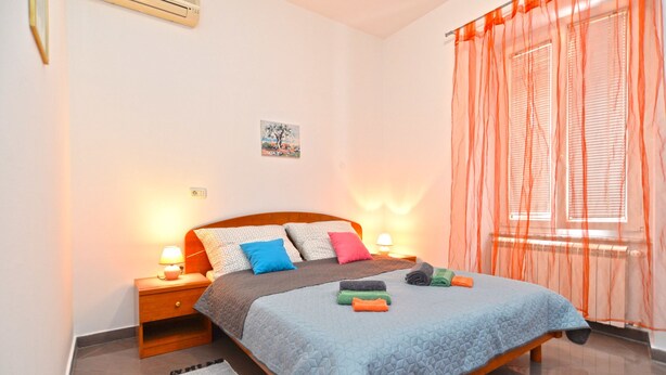 Une chambre à coucher comprenant un lit, des tables de chevet, un climatiseur fixé au mur, une fenêtre avec des rideaux légers et un radiateur.