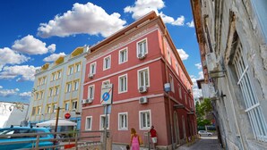 Une rue étroite en milieu urbain, avec un immeuble rouge, un immeuble bleu et un immeuble blanc. On y voit des voitures garées et un piéton traversant la rue.