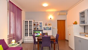 Un coin repas avec une table en bois et des chaises violettes, un meuble blanc avec des portes en verre et un téléviseur posé sur un meuble.