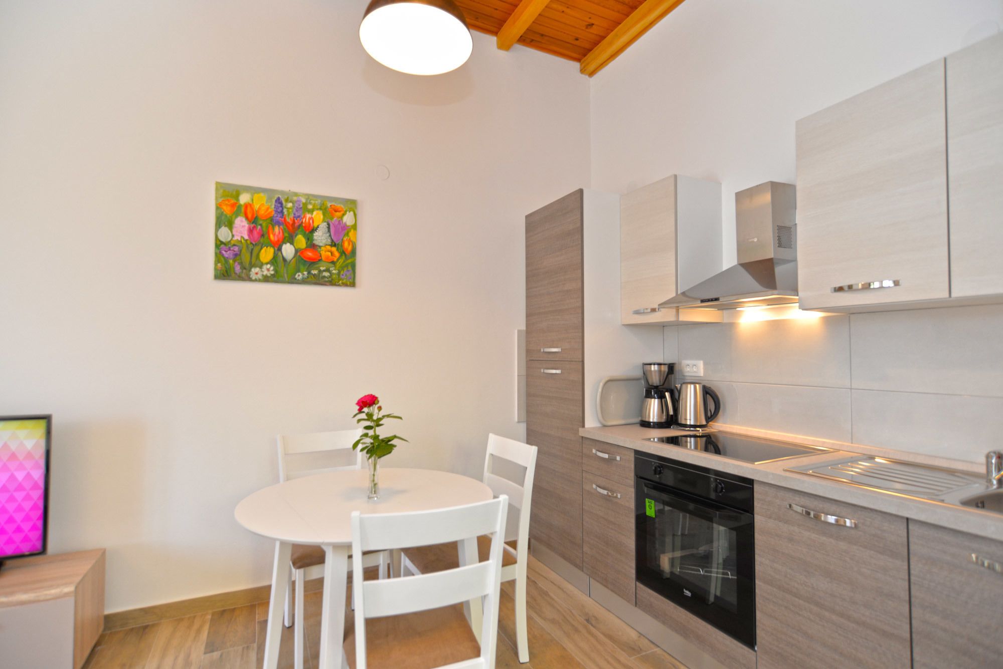A modern kitchen with a built-in oven, a coffee maker, and a sink. There is a dining table with chairs and a vase with a flower. A colorful painting hangs on the wall.