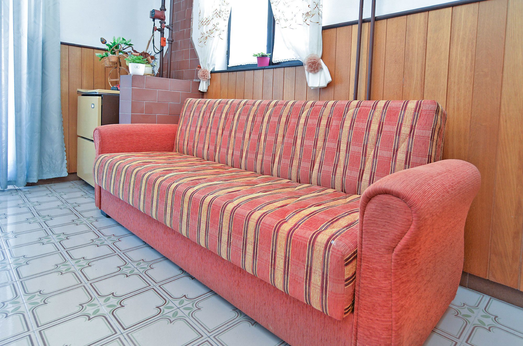 Ein rotes Sofa mit gestreiftem Kissen in einem Raum mit Fliesenboden, Holzvertäfelung und einem Fenster mit Vorhängen.