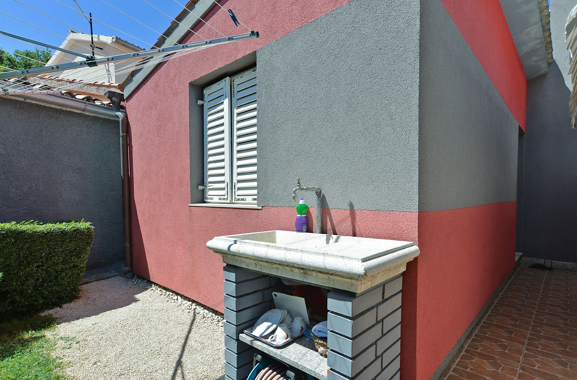 A small outdoor bathroom with a sink and window shutters, set against a red and grey wall.
