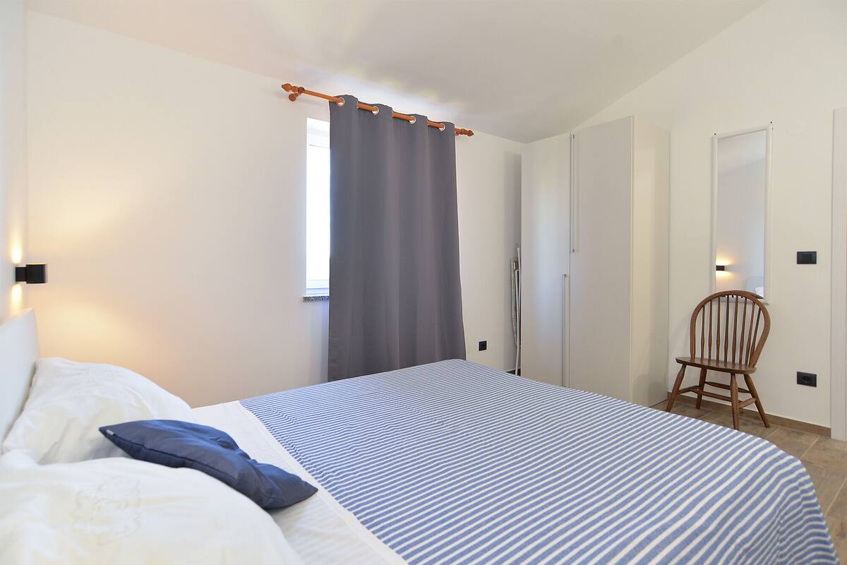 A bedroom with a bed, a chair, a window with curtains, and a wall-mounted light.