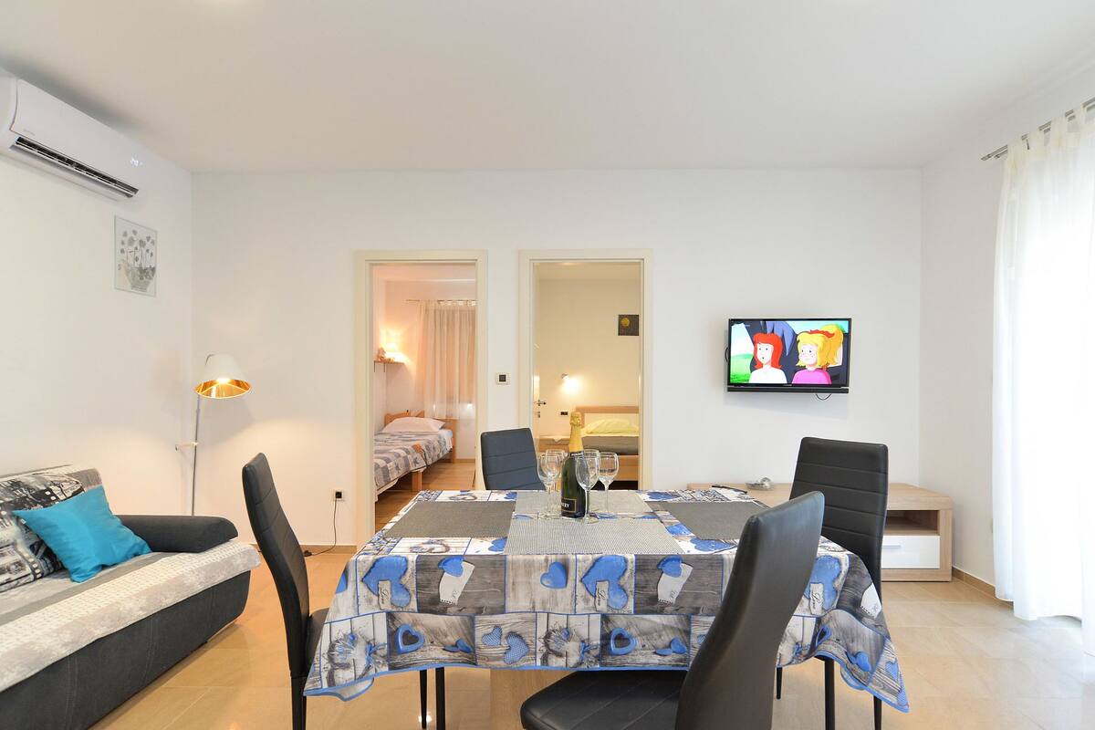 A modern living room with a sofa, a dining table with chairs, a TV, and a bedroom visible through an open door.