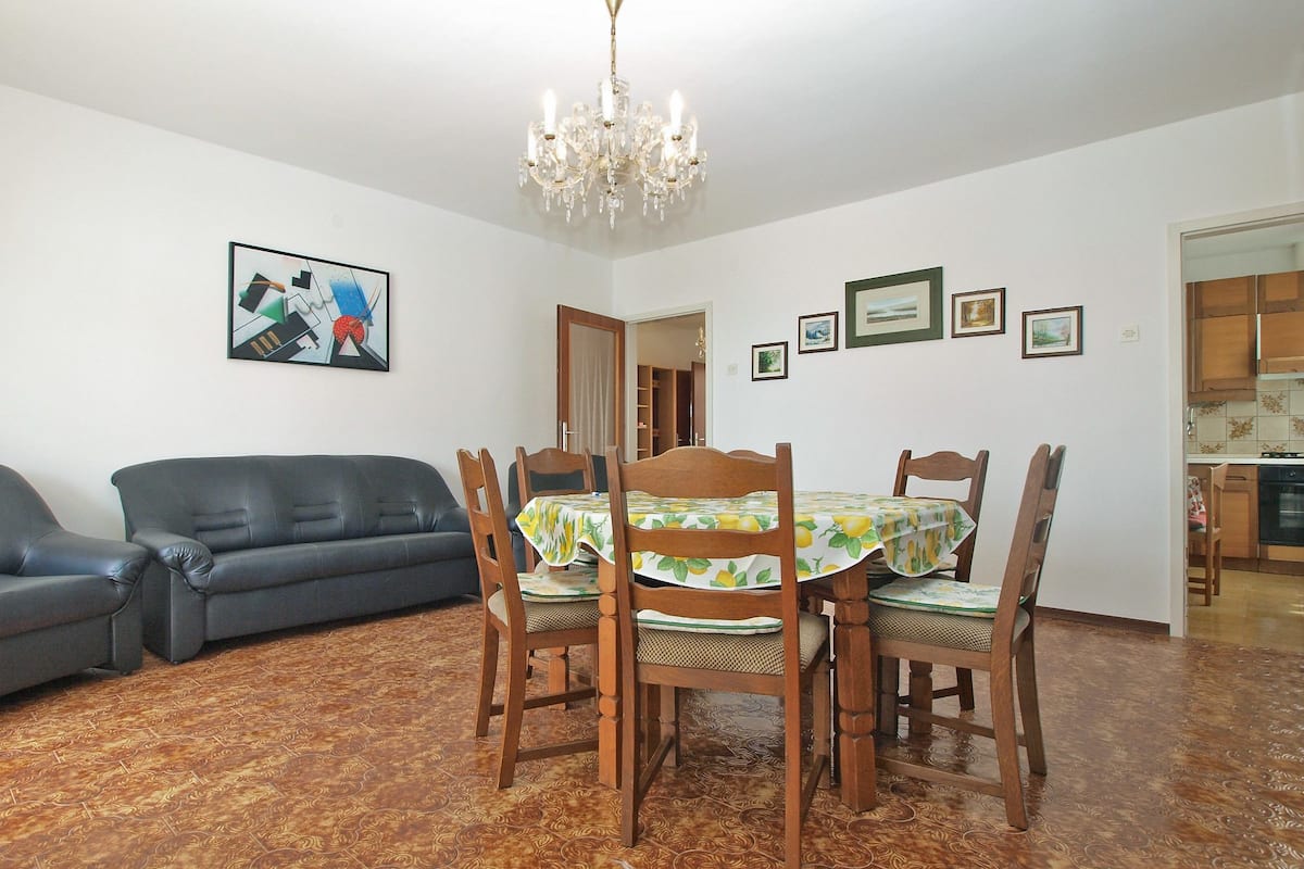 Un comedor con una mesa dispuesta para cuatro personas, rodeada de sillas de madera. Un candelabro cuelga del techo, y un sofá está ubicado contra la pared.