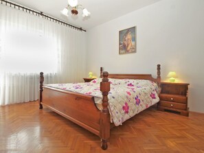 Une chambre à coucher avec un lit en bois, une literie à motifs floraux, deux tables de chevet avec des lampes, un tableau au mur et une fenêtre avec des rideaux légers.