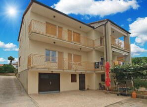 Un immeuble de deux étages doté d’un balcon à l’étage supérieur, d’un garage au rez-de-chaussée et d’un petit espace extérieur aménagé avec une table et des chaises.