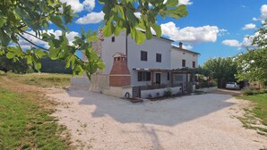 A two-story house with a stone chimney, a gravel driveway, and a car parked in the driveway. - House 1708 (Barban)