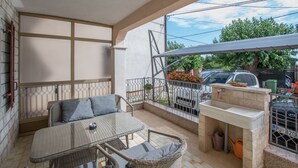 A balcony with a glass table and wicker chairs, a small barbecue area, and a view of parked cars and greenery. - Apartment 1709 (Umag)