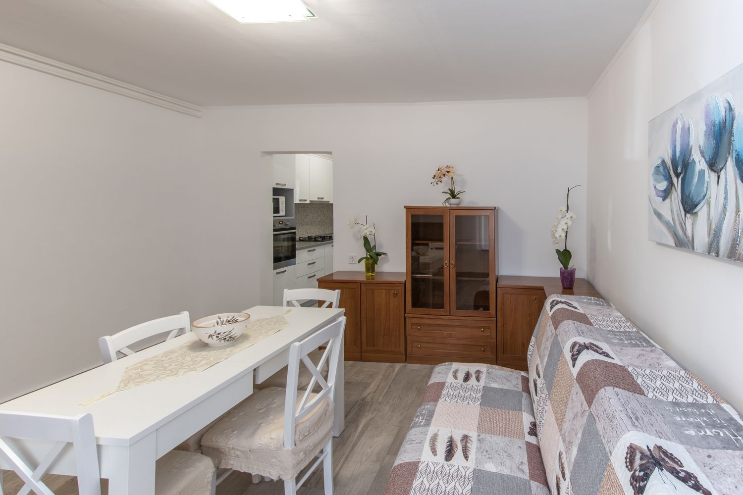 A compact living space with a dining table, white chairs, a sofa, a wooden cabinet, and a kitchen in the background.