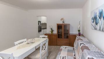 A compact living space with a dining table, white chairs, a sofa, a wooden cabinet, and a kitchen in the background.
