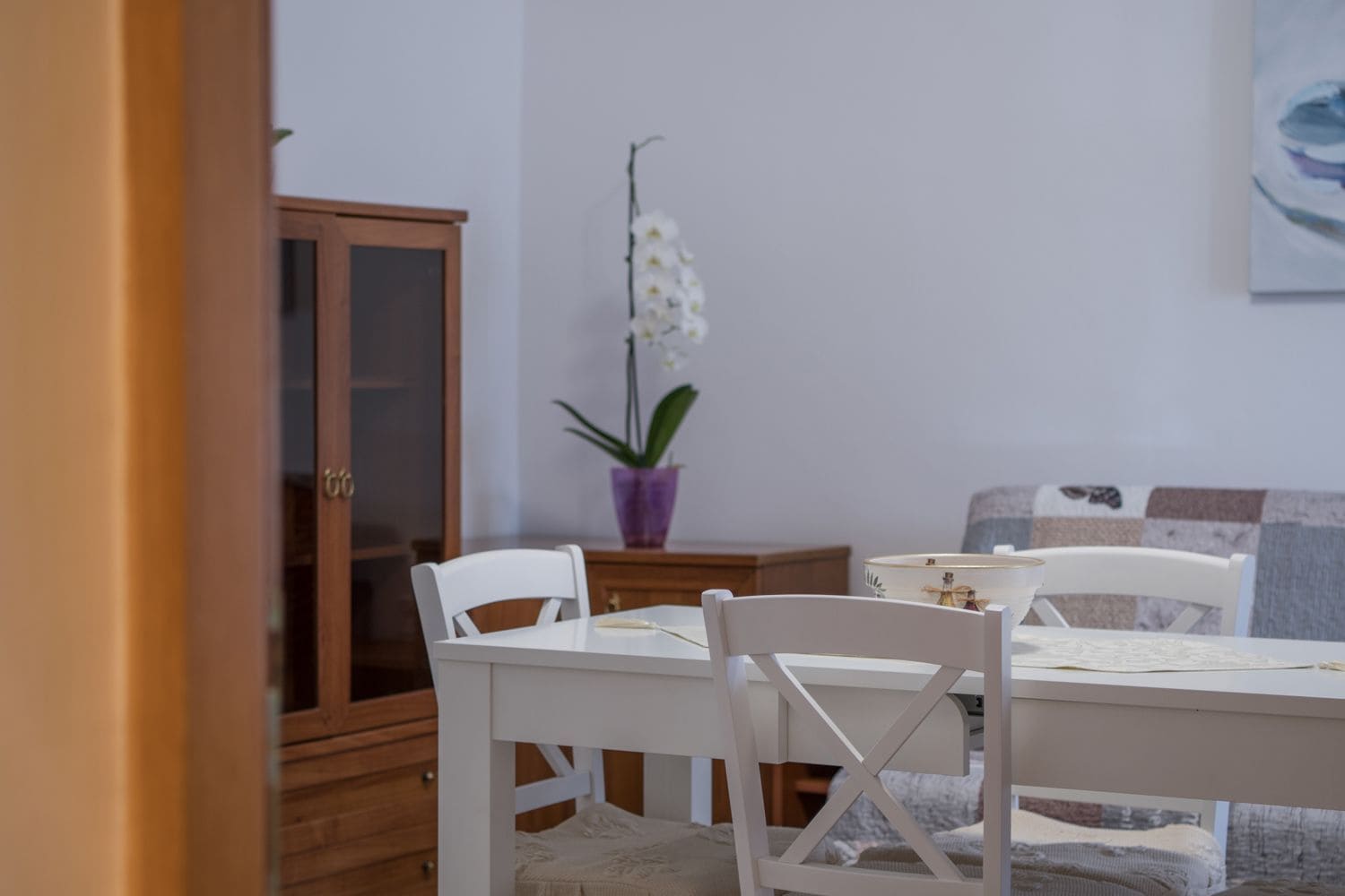 A dining area with a white table and chairs, a purple orchid plant, and a wooden cabinet.