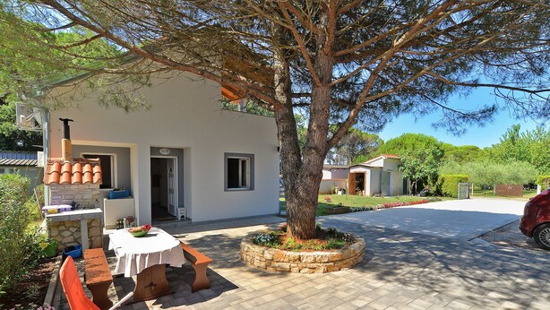 A white building with a stone bench and orange chairs outside, a tree in the center, and a paved driveway leading to a car. - Apartment 1713 (Fazana)