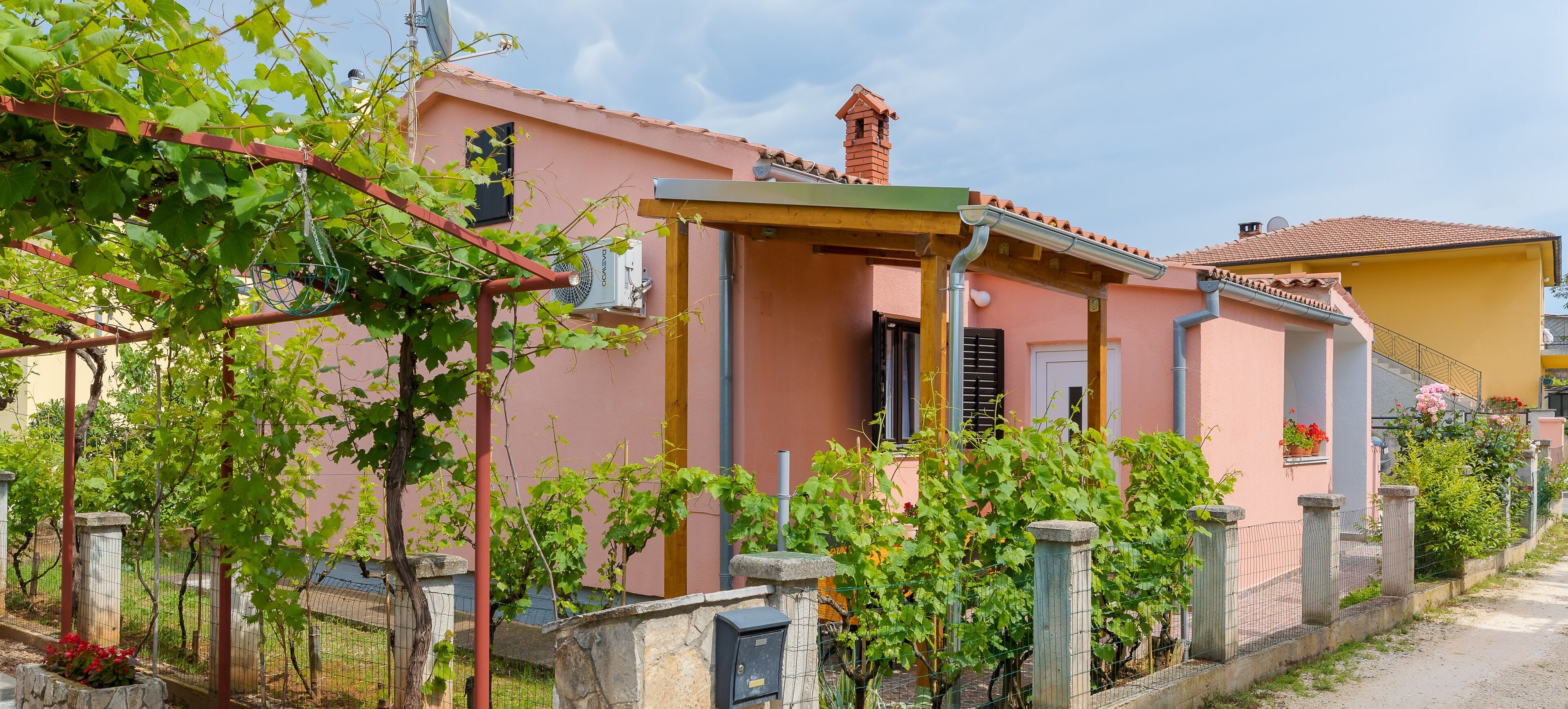 Une villa rose dotée d’un porche abrité, d’un appareil de climatisation visible et d’un petit jardin planté de vignes.