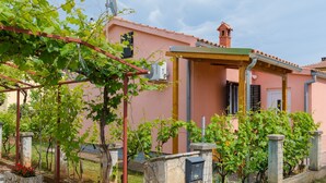 Una villa rosa con porche cubierto, unidad de aire acondicionado visible y un pequeño jardín con parras.