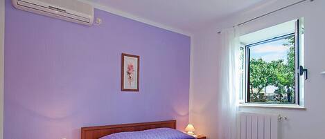 A bedroom with a bed, two bedside tables with lamps, a window with a view of greenery, and an air conditioning unit on the wall.