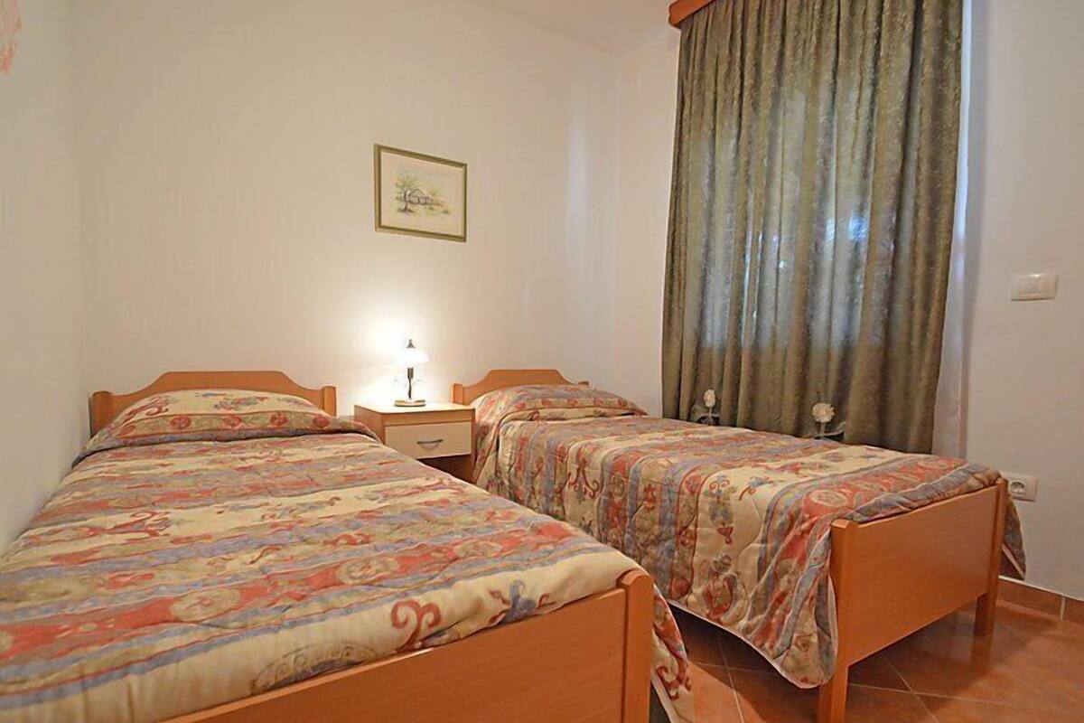 Une chambre à coucher avec deux lits en bois, une table de chevet, une lampe, un tableau encadré au mur et une fenêtre avec des rideaux.