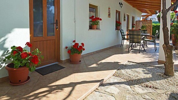 A covered outdoor area with a wooden door, potted red flowers, and a table with chairs. - Apartment 1770 (Kanfanar)