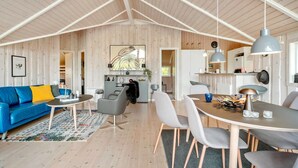Ein geräumiger Wohnbereich mit hoher Decke, einem blauen Sofa, einem Esstisch mit Stühlen und einer durchgehende Tür erkennbaren Küchenzeile.