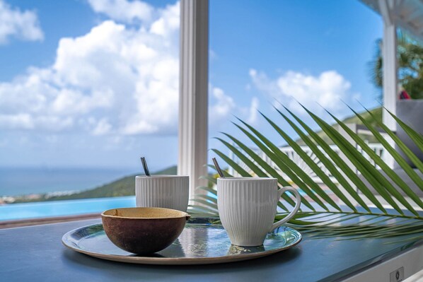 A table with a bowl and two cups, a palm leaf, and a view of the ocean and hills.