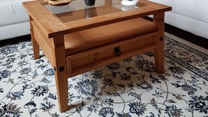 Una mesa de centro de madera con un jarrón y un cuenco sobre una superficie de vidrio, ubicada frente a un sofá blanco con alfombra estampada.