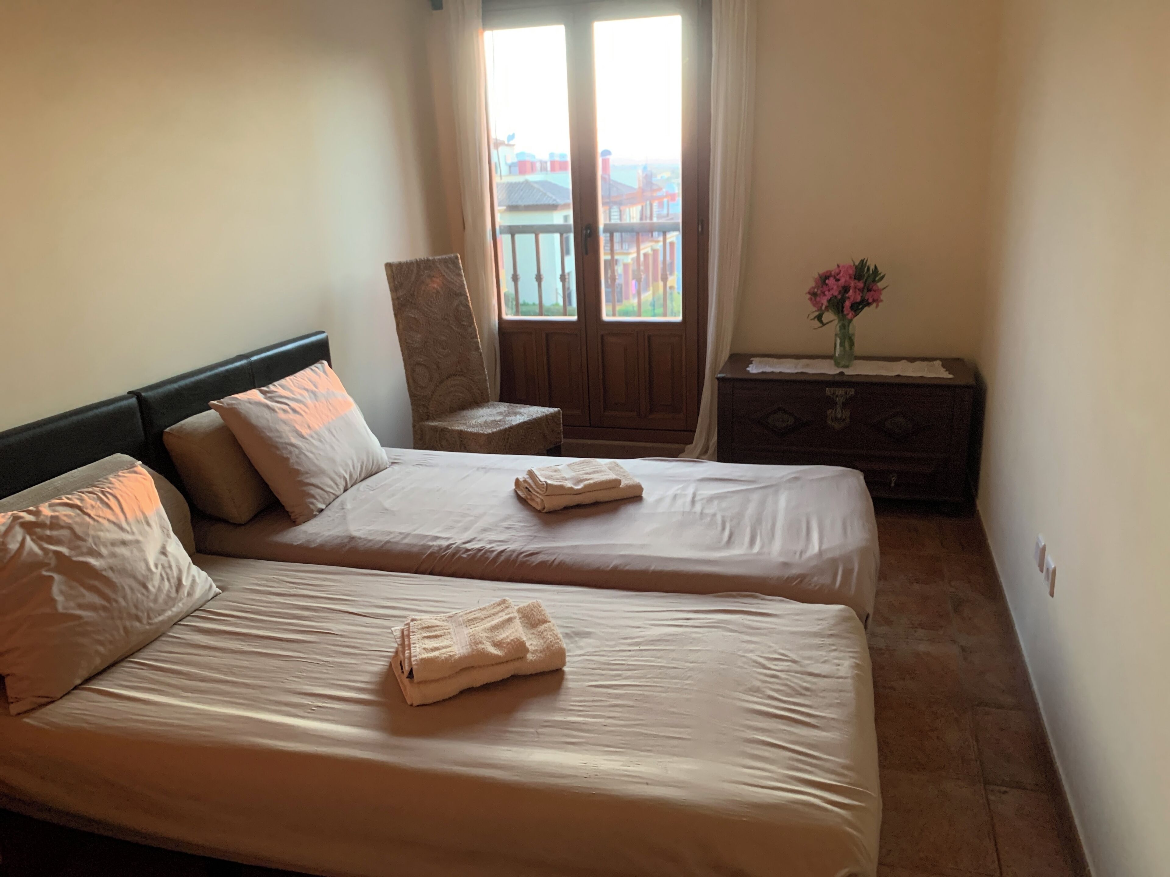 A bedroom with two beds, a wooden dresser, and a window with a view of buildings.