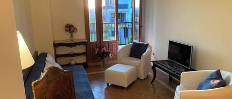A living room with a sofa, two armchairs, a coffee table, a TV, a lamp, a vase with flowers, and a view of a building through a glass door.